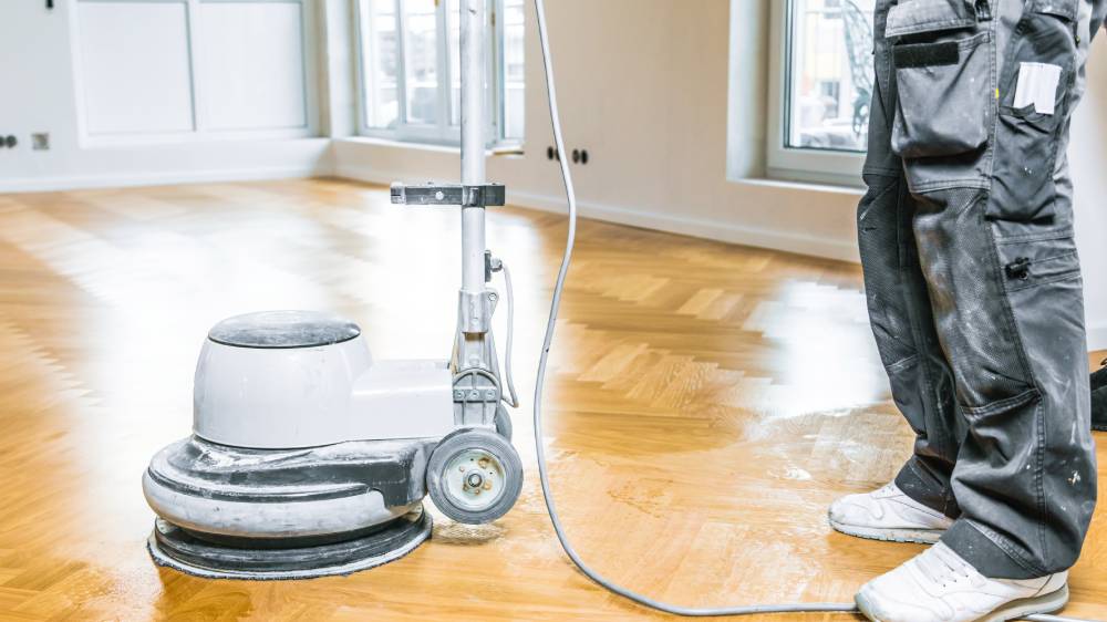 Flooring worker using a rotary floor sander to refinish a herringbone hardwood floor in a bright, empty Winnipeg home.