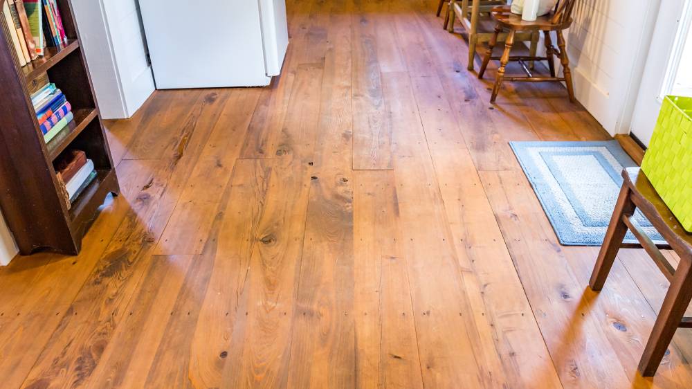 Refinished wide-plank hardwood floor with a warm honey finish and visible knots in an older Winnipeg home, with a wooden bookshelf, chairs and a blue rug.